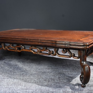 Pu&ograve; includere: Un tavolo in legno marrone scuro con intagli intricati sulle gambe e un piano rettangolare. Il tavolo &egrave; su una superficie grigia testurizzata.