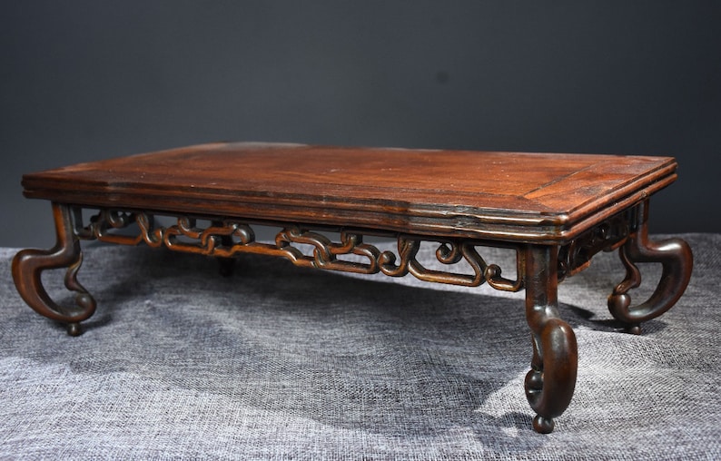 Pu&ograve; includere: Un tavolo in legno marrone scuro con intagli intricati sulle gambe e un piano rettangolare. Il tavolo &egrave; su una superficie grigia testurizzata.