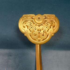 Op de afbeelding: Een goudkleurige metalen wandhaak met een decoratieve, waaiervormige bovenplaat met ingewikkelde, symmetrische krulmotieven en geometrische patronen. De haak heeft een gladde, gebogen arm die vanuit het midden van de kop naar beneden loopt.