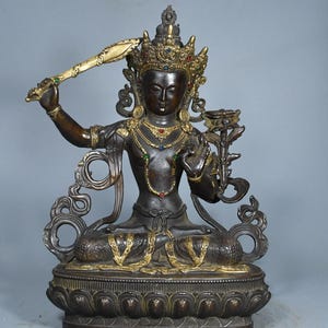 Op de afbeelding: Een donkerbronskleurig standbeeld van een zittende godheid met goudkleurige accenten, met een versierde kroon met rode en blauwe stenen. De figuur houdt een opgeheven zwaard in de rechterhand en een lotusbloem in de linkerhand op een lotusvoet.