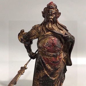 May include: A bronze-toned metal figurine of a warrior with a long beard, wearing ornate armor with red dragon detailing on the chest. The figure holds a long-handled polearm weapon in one hand and stands in a firm, upright posture.