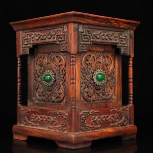 May include: A small, intricately carved wooden box with a square shape. The box is decorated with intricate floral patterns and has two green gemstone accents on the front. The box is made of dark wood and has a polished finish.