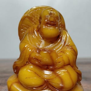 Peut inclure: Une statue de Bouddha de couleur ambre, richement détaillée. La figure assise affiche une expression sereine et est ornée d'un halo circulaire. La statue est sculptée avec des détails complexes, mettant en valeur les plis des robes et la posture du Bouddha.