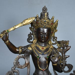 Op de afbeelding: Een donker bronskleurig standbeeld van een godheid met een versierde goudkleurige kroon en sieraden bezet met kleine rode, blauwe en groene stenen. De figuur houdt een opgeheven zwaard in de rechterhand en een lotusbloem met een boek in de linker.