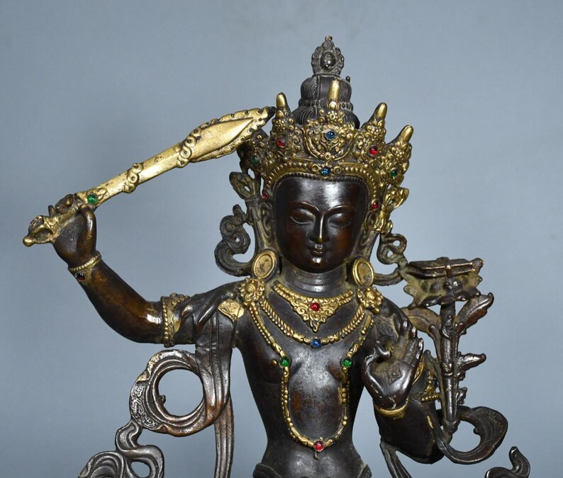 Op de afbeelding: Een donker bronskleurig standbeeld van een godheid met een versierde goudkleurige kroon en sieraden bezet met kleine rode, blauwe en groene stenen. De figuur houdt een opgeheven zwaard in de rechterhand en een lotusbloem met een boek in de linker.
