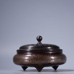 Op de afbeelding: Een bronzen wierookbrander met een deksel en drie poten. Het deksel heeft een kleine knop en een decoratief patroon. De behuizing van de brander heeft een ingewikkeld, zich herhalend ontwerp in een lichtere kleur. Het is een traditioneel Aziatisch decoratie-item.