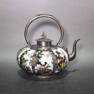 May include: A round white ceramic teapot featuring colorful painted figures and trees, encased in a silver-toned metal frame with a curved handle and a spout. The lid is topped with a small metal figure, and the body displays Chinese calligraphy.