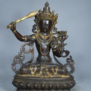 Op de afbeelding: Een donkerbronzen standbeeld van een zittende godheid met een versierde kroon ingelegd met rode, blauwe en groene stenen. De figuur houdt een opgeheven zwaard in de rechterhand en een lotusbloem in de linkerhand op een lotusvormige voet.