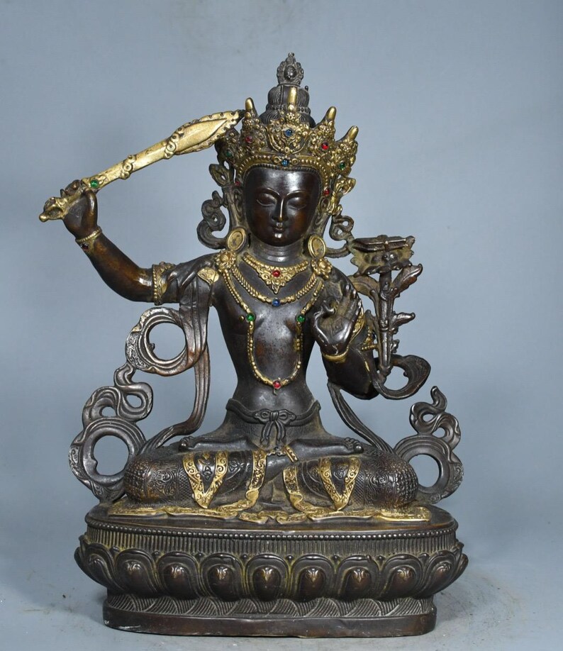 Op de afbeelding: Een donkerbronzen standbeeld van een zittende godheid met een versierde kroon ingelegd met rode, blauwe en groene stenen. De figuur houdt een opgeheven zwaard in de rechterhand en een lotusbloem in de linkerhand op een lotusvormige voet.