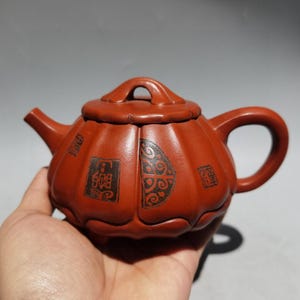 Puede incluir: Una tetera de arcilla de color marrón rojizo con cuerpo en forma de calabaza, asa curva y pico corto. La superficie presenta sellos rectangulares negros en relieve con caracteres chinos y un patrón decorativo semicircular en el lateral.