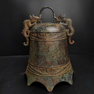May include: A bronze bell with a dark patina, featuring intricate dragon sculptures on either side of the handle. The bell's surface is covered in detailed inscriptions and decorative bands, showcasing an antique aesthetic.