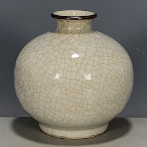 Könnte beinhalten: Eine cremefarbene, runde Keramikvase mit einem Craquelé-Glasurmuster. Die Vase hat einen kurzen Hals mit einem dunkelbraunen Rand und einem leicht ausgestellten Fuß. Die Gesamtform ist bauchig.