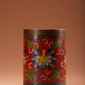 Op de afbeelding: Cilindrische decoratieve container met een rode achtergrond en bloemenontwerp. De container heeft een gedetailleerd patroon met blauwe, groene, gele en witte accenten. De boven- en onderranden hebben een bronzen afwerking, wat een vleugje elegantie toevoegt.