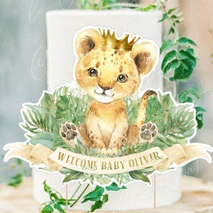 May include: A lion cub cake topper with a golden crown, surrounded by green leaves and a banner that reads "Welcome Baby Oliver".
