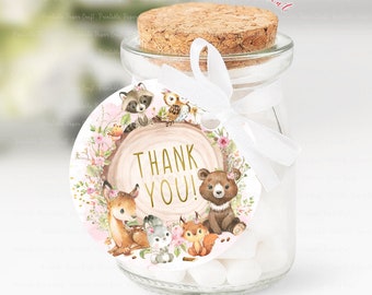 May include: Round thank you tag with a woodland theme. The tag features a brown wooden circle with the words "Thank You!" in gold lettering. The circle is surrounded by pink flowers, green leaves, and woodland animals including a deer, a bear, a raccoon, a squirrel, and an owl. The tag is attached to a clear glass jar with a cork lid and a white ribbon.