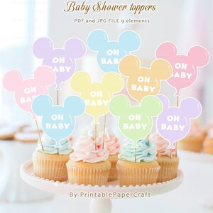 May include: Nine pastel-colored Mickey Mouse head shaped balloon cupcake toppers with the text "Oh Baby" in white. The toppers are on cupcakes with pastel-colored frosting.