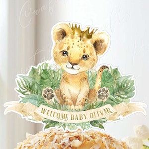 May include: A cake topper featuring a cute cartoon lion wearing a gold crown, surrounded by green leaves. The banner reads "Welcome Baby Oliver". The cake topper is labeled "Safari Animal - Cake Topper - By PrintablePaperCraft".
