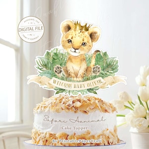 May include: A printable cake topper featuring a cute cartoon lion cub wearing a gold crown, surrounded by green leaves. The topper says "Welcome Baby Oliver" and "Safari Animal - Cake Topper - By PrintablePaperCraft".
