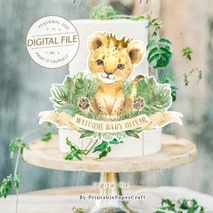 May include: Printable cake topper featuring a cartoon lion cub wearing a gold crown, sitting on green leaves with a banner that reads "Welcome Baby Oliver".