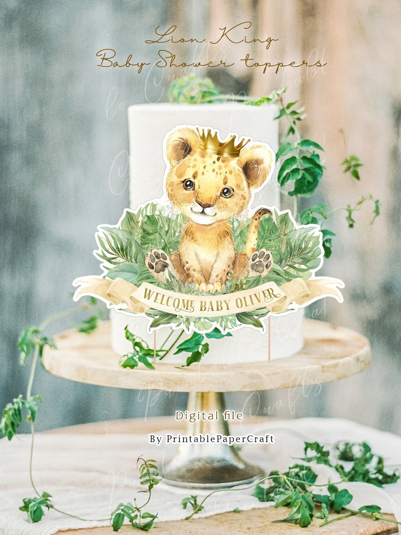 May include: A lion cub cake topper with a golden crown, surrounded by green leaves and a banner that reads "Welcome Baby Oliver".