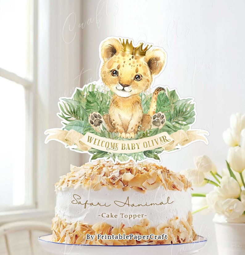 May include: A cake topper featuring a cute cartoon lion wearing a gold crown, surrounded by green leaves. The banner reads "Welcome Baby Oliver". The cake topper is labeled "Safari Animal - Cake Topper - By PrintablePaperCraft".