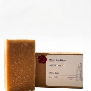 May include: A bar of artisan soap and its packaging. The soap is a light brown color and the packaging is a cardboard box with the words "Persimmon Artisan Soap" and "4 oz/113g" printed on it.