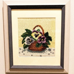 May include: A framed needlepoint embroidery of a basket of purple, yellow, and white pansies with green leaves.