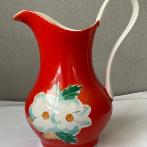 Peut inclure: Un pichet en céramique rouge avec une anse blanche et un motif floral. Les fleurs sont blanches avec des centres jaunes et des feuilles vertes.