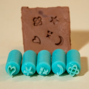 May include: Five turquoise rubber stamps with different designs: a heart, a star, a flower, a crescent moon, and a swirl. The stamps are arranged in a row on a light beige surface. A brown rectangular block with the same designs is in the background.