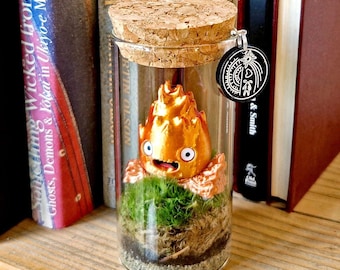 以下が含まれることがあります： コルクの蓋が付いたガラス瓶の中に、笑顔の小さな金色の置物が緑の苔に囲まれている。瓶は木の棚の上に置かれ、後ろには本が並んでいる。