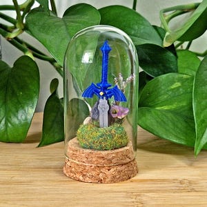 May include: A miniature blue sword with a winged crossguard is encased in a clear glass dome with a cork base. The sword is set into a patch of green moss, accompanied by small purple flowers and a dark stone, creating a detailed display piece.