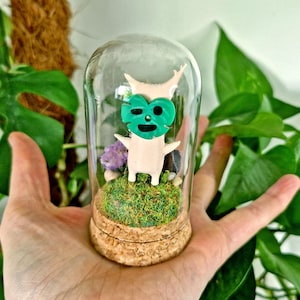 May include: A small, pale, humanoid figurine with a bright green monstera leaf mask stands inside a clear glass cloche. The figurine is positioned on a patch of green moss atop a circular cork base, surrounded by small decorative stones.