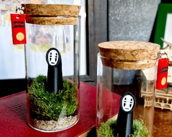 以下が含まれることがあります： コルクの蓋が付いたガラス瓶が2つあり、それぞれに映画「千と千尋の神隠し」の小さな黒いカオナシのフィギュアが入っており、緑の苔で囲まれています。瓶は赤い本の上に置かれています。