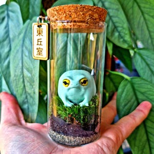 May include: A glass terrarium with a cork stopper, featuring a light blue frog figurine with yellow eyes, set on green moss. A small wooden tag with Japanese text is attached. The terrarium is held in a hand.