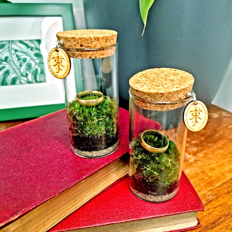 May include: Two glass terrariums with cork stoppers, each containing a gold ring atop green moss. The terrariums have wooden tags with a symbol. They sit on a red book, with a wooden table in the background.