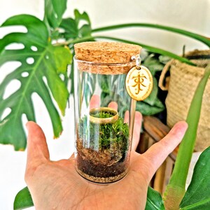 May include: A clear glass terrarium with a cork stopper, containing a miniature landscape with a gold ring, moss, and soil. A wooden tag with an inscription hangs from the top. The terrarium is held in a person's hand, with green foliage in the background.