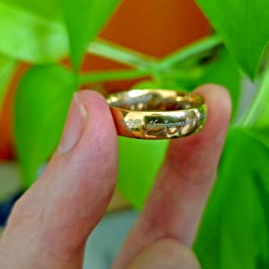 May include: A gold-colored ring with engraved text, held between fingers against a blurred green background. The ring's surface reflects light, highlighting the inscription. The ring is a piece of jewelry.