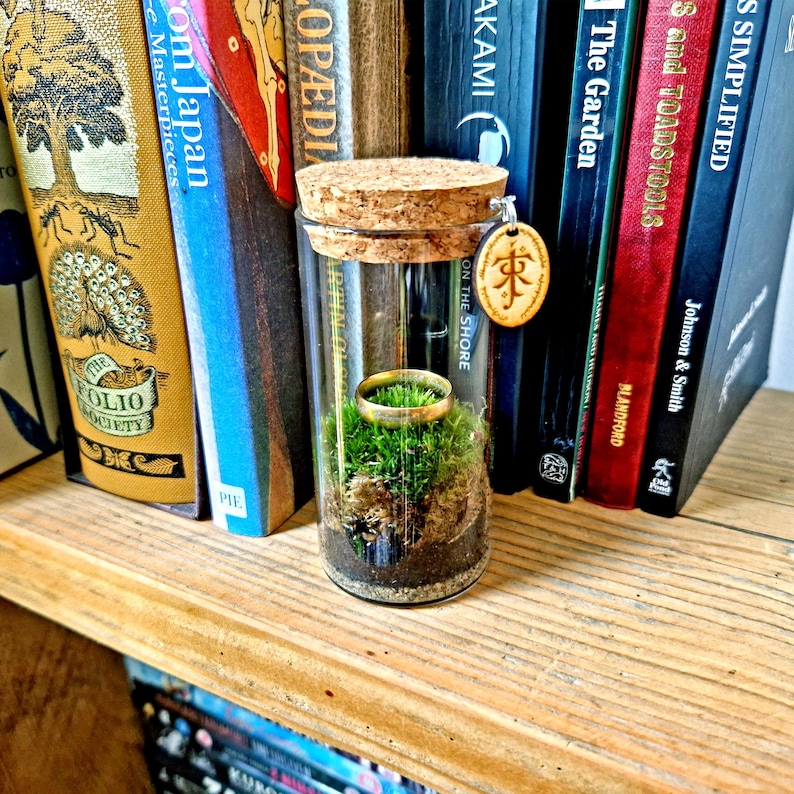 May include: A glass terrarium with a cork stopper, containing a gold ring atop green moss and soil. A wooden charm with an inscription hangs from the cork. The terrarium is displayed on a wooden shelf with a backdrop of various books.