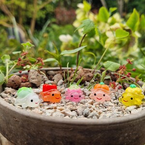 May include: A close-up of a small, round, grey ceramic pot filled with gravel and miniature, colourful turtle figurines. The turtles are green, orange, pink, and yellow, each with a smiling face. The background is blurred with greenery.
