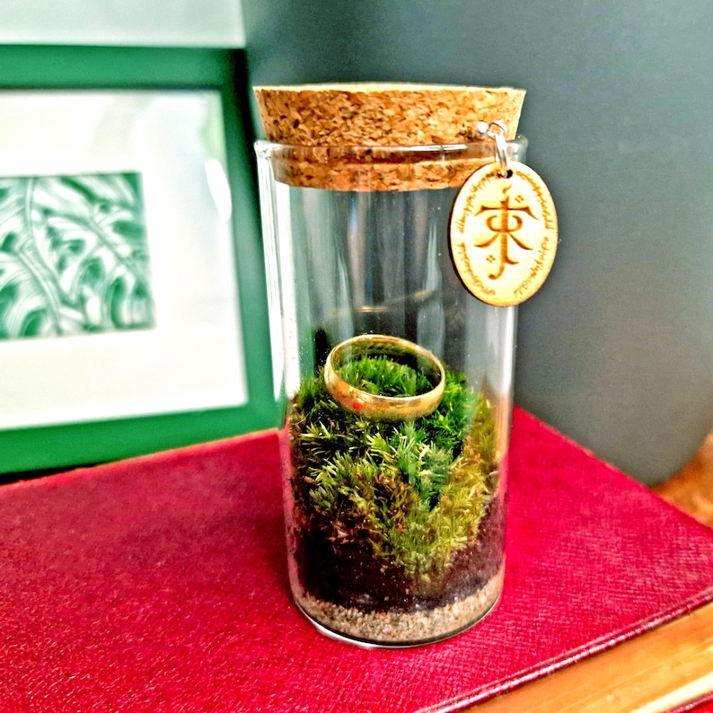 May include: A clear glass jar with a cork stopper, containing a gold ring resting on a bed of green moss. A small wooden tag with engraved text hangs from the jar. The jar sits on a red surface.