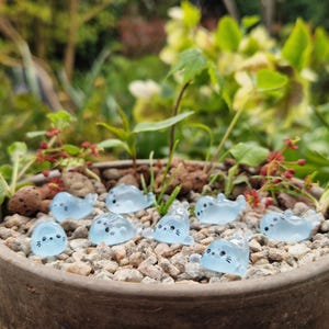 May include: A terracotta plant pot containing small, light blue seal figurines with black eyes and whiskers. The seals are on a bed of small, light-coloured pebbles, surrounded by green plants and red flowers.
