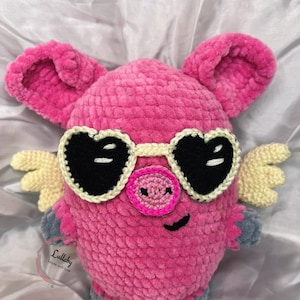 Puede incluir: Un cerdo rosa de crochet que lleva gafas de sol en forma de corazón con lentes negras y alas amarillas. El cerdo tiene una nariz rosa y una sonrisa cosida negra. El cerdo está sentado sobre una superficie blanca. El texto "Lullaby Made with love" es visible en un círculo blanco.