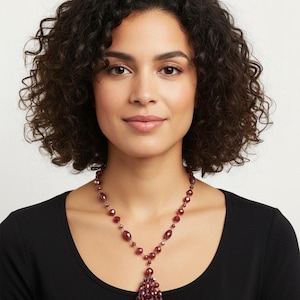 May include: A woman wearing a burgundy beaded necklace. The necklace features a series of small, round beads and larger, oval-shaped beads. A tassel of teardrop-shaped beads hangs from the center. The woman is wearing a black top.