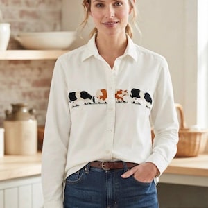 May include: A long-sleeved white button-down shirt featuring an embroidered horizontal row of four cows in black, white, and brown across the chest. The shirt has a classic collar and is tucked into dark blue denim jeans with a brown leather belt.