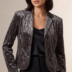May include: A charcoal gray crushed velvet blazer featuring a notched lapel collar, long sleeves, and a three-button front closure. The jacket is worn over a black satin camisole and black trousers, showcasing a textured, shimmering fabric finish.