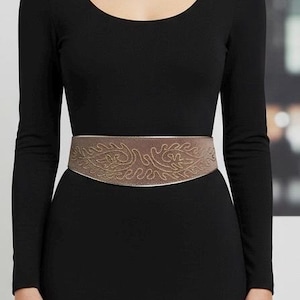 May include: A brown belt with an intricate, gold-colored embroidered design. The belt is worn over a long-sleeved black dress. The belt's design features a repeating pattern of swirling lines and leaf-like shapes.