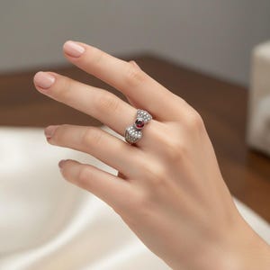May include: A silver-toned ring featuring a bow-shaped design adorned with small clear stones and a single round, deep red gemstone set in the center. The band is metallic and polished, showcasing a detailed construction with multiple sparkling accents.