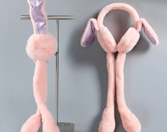 Könnte beinhalten: Rosa Plüsch-Hasenohrhut mit langen, baumelnden Beinen. Die Innenohren sind schimmernd lila. Der Hut hat ein Stirnband und Ohrenschützer. Der Text "Color: pink" befindet sich am unteren Rand des Bildes.