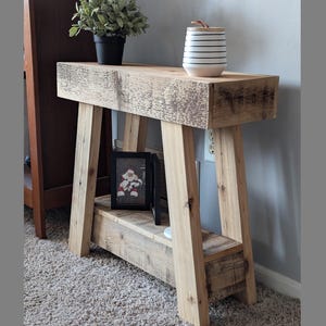 May include: A rustic wooden side table with a distressed finish. The table has a single shelf and a wide top. A small framed photo and a white ceramic jar with black stripes are on the table.