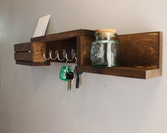 Può includere: Un porta chiavi in legno con uno scaffale e un piccolo cassetto. Il porta chiavi ha cinque ganci per appendere le chiavi. C'è un piccolo barattolo di vetro sullo scaffale.